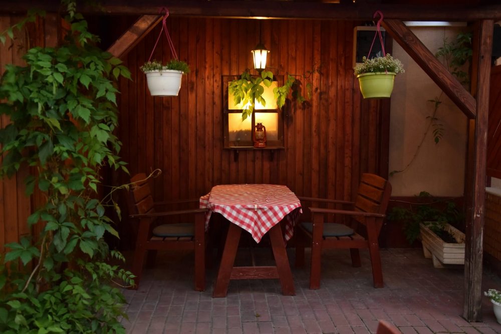 Faburkolatú kerti zug este, lámpással a falon, két paddal és egy asztallal, futtatott vadszőlővel